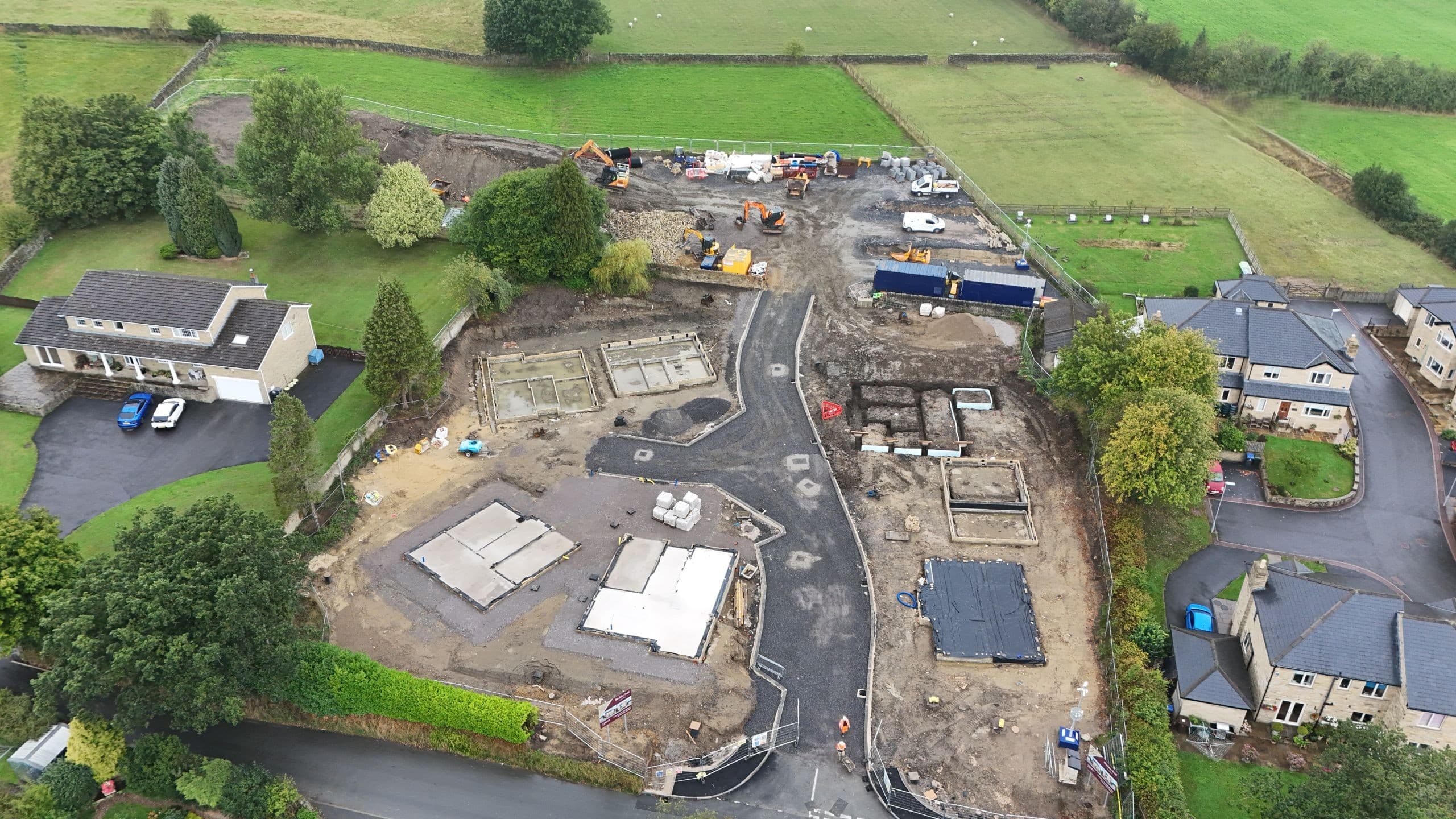 Aerial drone view of Birchley Fold, a new homes development in Silsden, showing early construction progress with foundations laid, a newly formed access road, construction machinery and site cabins, surrounded by green fields, mature trees and neighbouring homes.